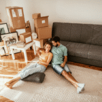 A couple sitting on the floor of a new apartment with moving boxes behind them looking happy