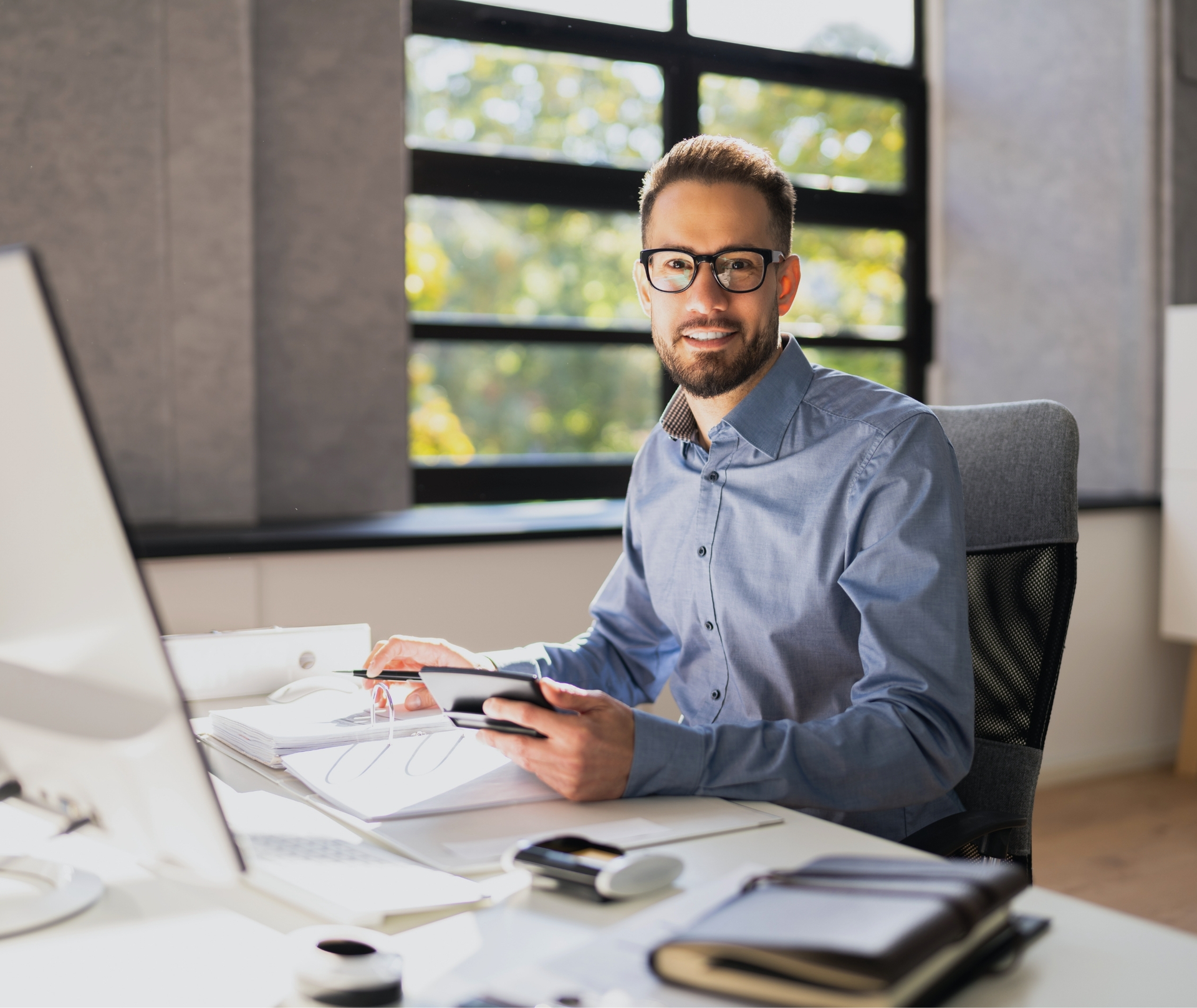 A male accountant/business owner sitting at his desk. There is a window behind him and natural light.