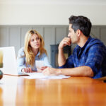 A woman and man in their 30s-40s are sitting at a dining table with laptop open, discussing interest rates and the Australian property market
