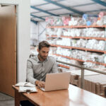 A business owner sitting at his desk working on laptop with stock shelves in the background