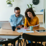 Stressed man and woman talking about family budget and savings, calculating household bills and expenses while sitting at table together with laptop computer, digital tablet and two cups of coffee.